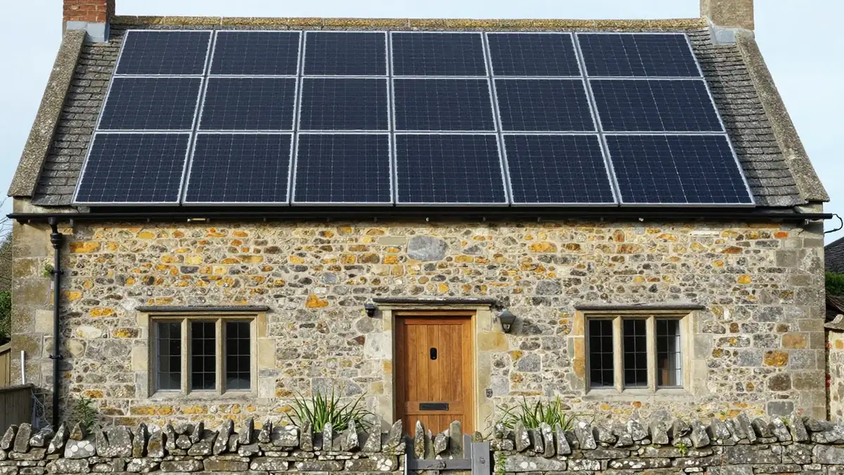 Stone cottage with a slate roof carrying two rows of black solar panels, a wooden front door and a dry-stone garden wall